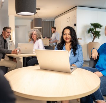 Several people in a modern office meet in a relaxed atmosphere at round tables. Two women in the foreground are working on laptops, while others in the background are engaged in conversation.