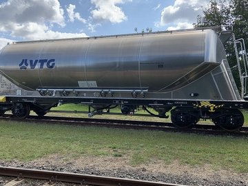 A stainless steel tank wagon with a VTG logo is stationed on the tracks, surrounded by grassy ground and trees under a partly cloudy sky.