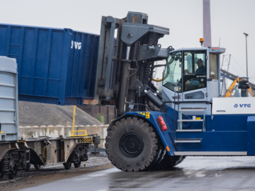  A large forklift truck lifts a VTG container onto a freight wagon. The scene shows a loading process in damp weather.